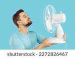 Small photo of Man using an electric fan to survive the summer heat. Happy guy standing with his eyes closed on a blue background, holding a white fan in his hands, and enjoying cool, fresh air blowing in his face