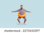 Small photo of Funny fat man in retro swimsuit and sunglasses poses with swim tube around his waist isolated on blue background. Happy plump bearded man with swim ring having fun and enjoying summer holiday at beach