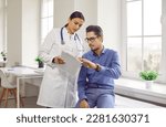 Doctor and patient talking during medical checkup at clinic. Young woman in white coat showing and explaining analysis results to man sitting on medical couch in examination room
