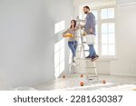 Small photo of Happy young couple painting the wall of their new home holding paint rollers and standing on the ladder. Married man and woman doing repair renovation preparing to move into a new flat.