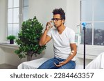 Small photo of Young African American man drinking glass of water while sitting on medical bed at clinic and receiving modern intravenous anti stress vitamin therapy through sterile IV drip line infusion system