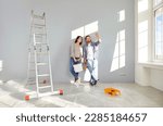 Young family couple doing renovations at home. Happy man and woman painting walls and decorating their house. Husband and wife with bucket and roller standing by wall and discussing interior design