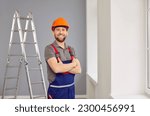 Small photo of Portrait of a construction worker or repairman in the house. Happy repair man wearing a safety helmet and workwear overalls standing with folded arms in the room, with a stepladder in the background