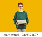 Small photo of Funny redhaired schoolboy with glasses with droll surprised expression face looks at the camera, dressed in green sweater and gray jeans, holding several school notebooks on isolated yellow background