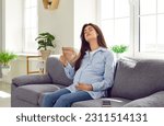 Small photo of Pregnant mother woman in third trimester expects baby, suffers from extreme summer heat and hot flush hormone change symptoms, sits on sofa in too stuffy overheated room, and refreshes with hand fan