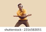 Small photo of Positive cool dark-skinned man rejoices, has fun and dances isolated on background of mocha color. Funny african american man in casual clothes and sunglasses dancing waving his arms and laughing.