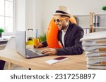 Small photo of Funny happy business man sitting at the desk of his workplace in office with juice cocktail, boarding pass and passport holding rubber ring and booking tickets for summer vacation online via laptop.