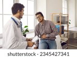 Small photo of Young guy with acute pain in right side talking to doctor. Adult ethnic patient with stomachache, kidney bloating or cirrhosis asking for help during consultation appointment in exam room at hospital