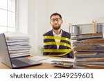Small photo of Funny man, stuck in office like work slave, bound with yellow adhesive tape sitting at desk with notebook computer and piles of paper folders and binders and looking at camera with sad face expression