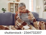 Sick senior woman checking her temperature wrapped in a blanket on the sofa suffering from headache. Elderly person with seasonal flu or cold feel unhealthy with influenza at home.