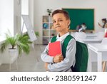 Small photo of Proud boy first grader with satchel and workbook posing on first day of elementary school getting ready to receive quality assignment standing in classroom with big blackboard. Back to school