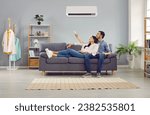Small photo of Young family couple enjoying life in their apartment with a modern air conditioning system. Happy husband and wife relaxing on the sofa at home and setting up the temperature on their AC on the wall
