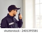 Small photo of Male security guard using portable radio transmitter indoors. Security guard wearing sunglasses, black uniform and cap standing in front of window using walkie-talkie radio
