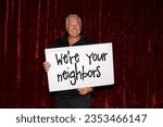 Small photo of Photo Booth. Large Sign. A man smiles and laughs as he holds a large sign that reads We're your neighbors while in a Photo Booth. Neighbors love a Photo Booth for all parties and events