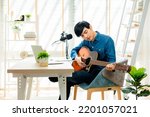 Small photo of Asian young man songwriter play acoustic guitar listen song from smartphone think and write notes lyrics song in paper sit in living room at home studio. Music production at home concept.