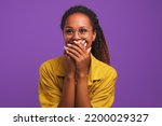 Small photo of Young laughing pretty girl African American woman covers mouth with hands tries to hide joy or laughter after hearing joke dressed in yellow shirt stands in purple solid background