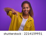Young horny beautiful African American woman in casual clothes pointing down to draw your attention to important message or to tell about crazy discounts stands on plain purple background. Copy space