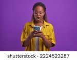 Small photo of Young shocked African American woman mouth wide open looking at screen of mobile phone surprised by large bill for payment or very good promotional offer stands on lilac studio background