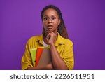 Young intrigued pretty African American woman student holds workbooks looks at camera with frightened face, opening mouth in surprise or fear stands on purple background. Education, training, exams