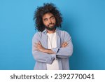 Small photo of Young doubtful funny Arabian man rolls eyes and looks up with offense crossing arms in front of chest after insult or inappropriate remark from boss posing on isolated blue background.