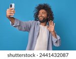 Small photo of Young affable excited Arabian man makes video call through mobile phone and says hello waving hand rejoicing in long-awaited conversation with friend from college stands posing in blue studio.