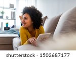 Social media time. Staying in touch with her tablet. Cheerful woman sitting on couch using tablet pc at home in the living room. Smiling young woman with tablet on the sofa