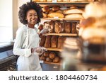 Smiling baker woman standing with fresh bread at bakery. Happy african woman standing in her bake shop and looking at camera. Satisfied baker with breads in background. Smiling woman at bakery shop