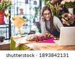 Smiling Woman Florist Small Business Flower Shop Owner. She is using her telephone and laptop to take orders for her store. Female gardener noting client order during mobile phone conversation