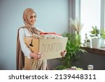 Small photo of Muslim woman with donation box at home. Donation box for poor with clothing in female hands. Woman donates clothing to shelter. Cheerful woman holds box of donated clothing.