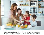Small photo of Family of four at the table, mother serves them breakfast. Everyone is happy and smiling, parents enjoy spending time with their children. A cheerful family has fun during a meal at the dining table.