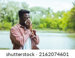 Small photo of Sick Man With High Temperature is Using Handkerchiefs, Trying to Override the Sickness While Walking in Nature. Allergic black man blowing on wipe in a park on spring season