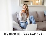 Woman drinking wine while using laptop. Young female is sitting at sofa. She is wearing striped t-shirt at home. Brunette sitting on couch in living room, drinking wine and using laptop.