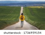 Long journey. Hiking woman walking on empty road. Hiker with hat and backpack wearing yellow waterproof jacket. Travel concept. Getting away from it all