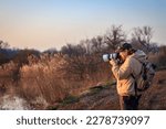 Landscape photographer with camera and telephoto lens photographing wildlife in nature during sunset