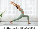 Side view of Asian woman wearing green sportwear doing Yoga exercise in front of windows,Yoga high lunge pose or Anjaneyasana,Calm of healthy young woman breathing and meditation yoga at home