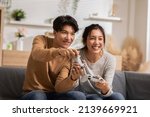 Asian young couple sitting on couch in living room enjoy playing video games spending time together. Happy couple play video games at home both are smiling, laughing and enjoying moment on weekend.
