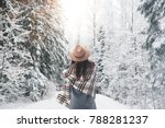 Beautiful woman standing among snowy trees in winter forest and enjoying first snow. Wearing hat, plaid scarf and coat. Wanderlust and boho style