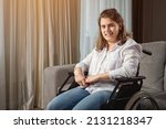 Small photo of Young woman with infantile cerebral paralysis in white shirt smiles sitting in wheelchair near stylish sofa in living room at home, sunlight.