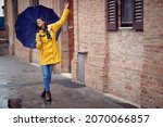 Small photo of A young handsome woman in a yellow raincoat and with umbrella is dancing while walking the city in a relaxed manner on a rainy day. Walk, rain, city
