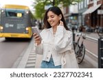 Small photo of Happy commute on public transport. Multi-ethnic attractive woman with beautiful smile at bus station looking at phone
