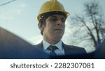 Small photo of A Young, Beardless Construction Engineer or Contractor in a Helmet and Suit Examining Blueprints in a Blue Folder and Getting Frustrated over a Faulty Detail
