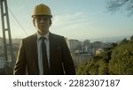 Small photo of Handsome, Suit-and-Tie-Clad, Beardless, Helmeted Businessman or Urban Planner Smiling at the Camera with a Cityscape Background