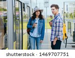 Lifestyle photo of young woman and man holds with cup of coffee waits while opening door to tram. Portrait of female traveler In denim jacket with backpack talking with handsome guy on tram stop.