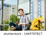 Lifestyle portrait of hipster student man with smart phone sitting on bench with yellow bagpack and phone on the tram stop in the modern city.