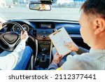 Man instructor teaching student about traffic rules on first automobile driving class. Examiner sitting with her student inside a car. Writing points. Driving instructor writing points