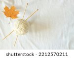 White wool thread ball with wooden bamboo knitting needles and autumn maple fall leaf on white linen fabric background. Top view, copy space. Hobby, relaxation, mental health, sustainable lifestyle
