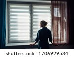 Small photo of Woman's silhouette at window in the background light. Young woman in gown stands near window with blinds. Quarantine, self-isolation, stay home, self-preservation, coronavirus pandemic.
