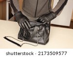 Robber in black outfit and gloves see on opened stolen women bag. A thief evaluates the value of stolen items from a womans handbag in kitchen interior