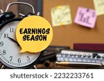 Business concept. The alarm clocks have a sticker with the inscription - Earnings Season. There are office items in the background in a blurry background.
