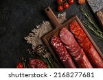 Small photo of Sausage and cold cuts. Types of different loaf sausages on an old wooden board on black background and burlap with cherry tomatoes, rosemary, garlic and spices.Rustic.Top. Background image, copy space